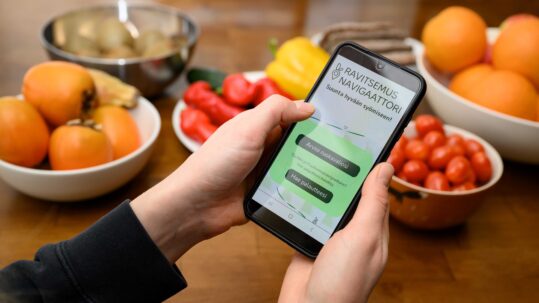 A phone being hand held and showing the ravitsemusnavigaattori website. In the background there are some vegetables held in plates.